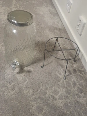 Glasbehållare med tappkran och ställning - Stor glasbehållare med mönstrad yta, silverfärgad metallkran och lock. Tillhörande svart metallställning med tre ben. Perfekt för att servera dryck på fester eller picknick. Rymmer flera liter och är enkel att använda tack vare tappkranen.