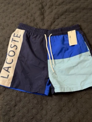 Flerfärgade badbyxor från Lacoste - Snygga badbyxor från Lacoste i färgblock med mörkblå, blå och ljusblå paneler. Stort Lacoste-tryck på ena benet och dragsko i midjan. Bakficka med broderad krokodil-logga. Perfekta för stranden eller poolen. STORLEK XL, PASSAR M. HELT NY, OANVÄND.