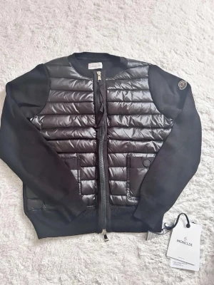 Svart cardiganjacka från Moncler - Snygg svart cardiganjacka från Moncler med quiltad, glansig front och stickade ärmar. Jackan har rund halsringning, dragkedja framtill och två fickor med knapp. Moncler-logga på ärmen och exklusiv känsla i materialmix av dun och ull.