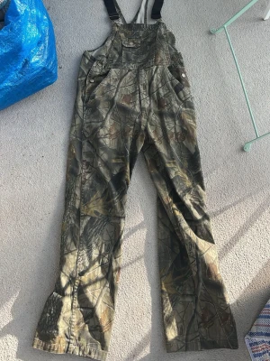Camouflage byxdress från Outfitters Ridge - Säljer en camouflagefärgad byxdress från Outfitters Ridge i 100% bomull. Byxdressen har klassiska hängslen, flera fickor framtill och vida ben. Perfekt för dig som gillar en avslappnad och cool stil.