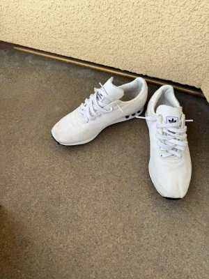 Vita Adidas sneakers med mesh - Fräscha vita sneakers från Adidas med klassisk logga på plösen och ventilerande meshmaterial. Skorna har vita snören och diskreta svarta detaljer på sidan. Perfekta för en sportig och stilren look.