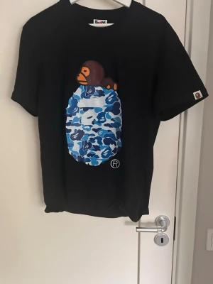 Svart BAPE t-shirt med blått camo - Svart t-shirt från BAPE med ikoniskt tryck av en apa ovanpå ett blått camoägg på bröstet. Klassisk passform och rund hals. T-shirten har korta ärmar och är tillverkad i mjuk bomull. Perfekt för dig som gillar streetwear och unika prints.