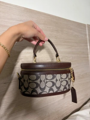 Coach brun handväska med monogram - Säljer en oval handväska från Coach i brunt och beige med klassiskt monogrammönster. Väskan har guldfärgade detaljer, dragkedja och ett kort handtag upptill. Perfekt accessoar för att lyfta din outfit. Den har. En lång kedja med , den är lite seg att stänga men det går utan problem