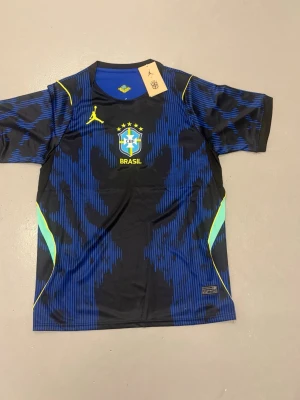 Brasiliens vm tröja - Snygg blå och svart Brasilien fotbollströja från Jordan med gula detaljer och Brasilien-logga på bröstet. Tröjan har korta ärmar, rund hals och är tillverkad i ett lätt och ventilerande material som passar perfekt till träning eller match.