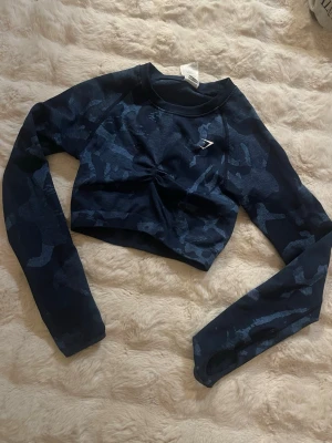 Mörkblå camo crop top från Gymshark - Snygg mörkblå långärmad crop top från Gymshark med camo-mönster och diskret logga på bröstet. Tröjan har åtsittande passform och rynkdetalj framtill. Perfekt för träning eller gymmet.