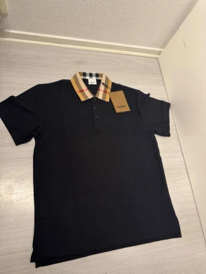 Svart Burberry piké med rutig krage - Svart skjorta från Burberry med korta ärmar och klassisk rutig krage i beige, svart, vitt och rött. Piké har knappar framtill och är tillverkad i mjuk bomull. Perfekt för dig som gillar stilrena detaljer och ikonisk design. Helt oanvänd. Storlek L. Fraktar samma dag eller dagen efter.  :)