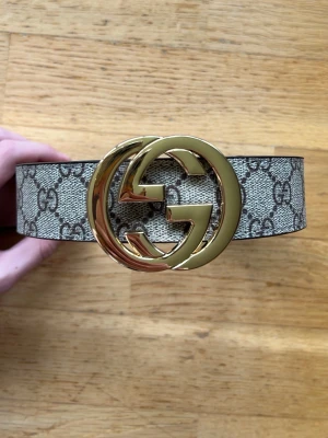 GG Bälte - Snyggt GG bälte med klassiskt GG Supreme-mönster i beige. Bältet har ett stort, glänsande guldspänne med dubbla G-loggan. Perfekt accessoar för att lyfta vilken outfit som helst.
