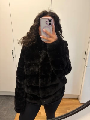 Svart fluffig fuskpälsjacka - Säljer en svart, supermjuk och fluffig fuskpälsjacka med hög krage och breda paneler. Jackan har en avslappnad passform och stängs med knappar framtill. Perfekt för dig som vill ha en varm och trendig look under vintern.
