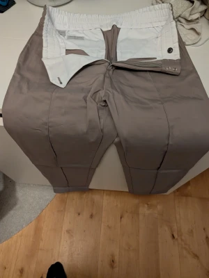 Beige kostymbyxor Tiger of Sweden - Snygga beige kostymbyxor från Tiger of Sweden med elastisk midja och klassisk passform. Byxorna har dragkedja och knäppning fram samt markerade pressveck längs benen. Perfekta för en stilren och modern look.