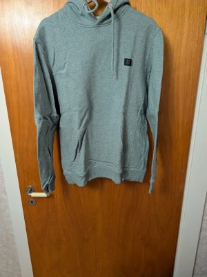 Grå hoodie från Les Deux - Snygg grå hoodie från Les Deux med broderad logga på bröstet och klassisk känguruficka. Mjuk bomullsblandning och justerbar dragsko i huvan. Perfekt för en chill och avslappnad stil.