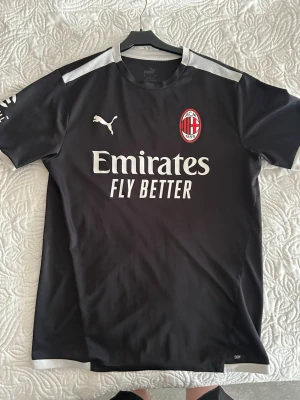 Svart AC Milan träningströja Puma - Snygg svart träningströja från Puma med AC Milan-logga på bröstet och vit text 'Emirates Fly Better' framtill. Tröjan har vita detaljer på axlarna och är tillverkad i ett lätt och ventilerande material, perfekt för fotbollsträning eller gym.