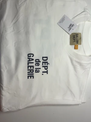 Vit t-shirt från Gallery Dept. - Vit t-shirt från Gallery Dept. med trycket 'DÉPT. de la GALERIE' i svart på framsidan. Klassisk rund halsringning och korta ärmar. Perfekt för en stilren och trendig look.