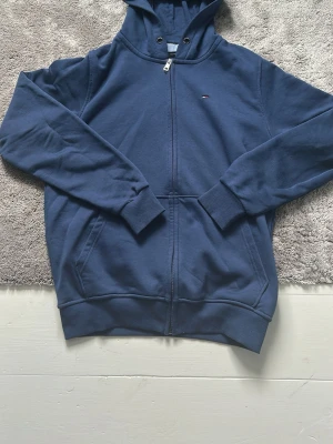 Blå zip hoodie från Tommy Hilfiger - Mörkblå hoodie med dragkedja från Tommy Hilfiger. Klassisk modell med huva, ribbade muddar och fickor framtill. Liten broderad logga på bröstet. Skön bomullsblandning som passar perfekt till chill dagar.