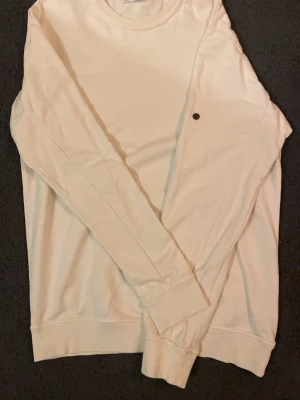 Vit långärmad sweatshirt - En stilren vit långärmad sweatshirt med rund hals och ribbade muddar. Tröjan har en liten svart detalj på bröstet och är perfekt för en avslappnad look. Passar bra till jeans eller mjukisbyxor.
