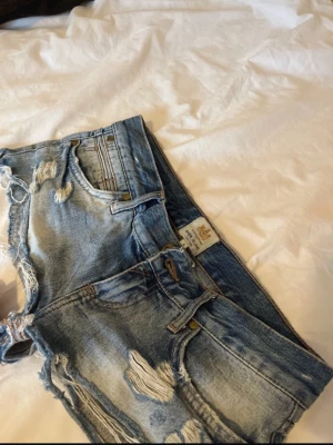 Blå slitna jeansshorts River Island - Säljer ett par blå jeansshorts från River Island med slitna detaljer och råa kanter. Shortsen har klassisk femficksdesign, bälteshällor och knappstängning. Perfekta för en avslappnad och trendig look.