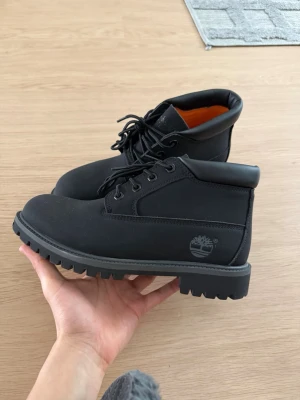 Svarta Timberland stövletter - Säljer ett par svarta stövletter från Timberland med vattentätt material och robust sula. Skorna har snörning, diskret logga på sidan och en klassisk, stilren design. Insidan är orange och materialet känns slitstarkt och tåligt.