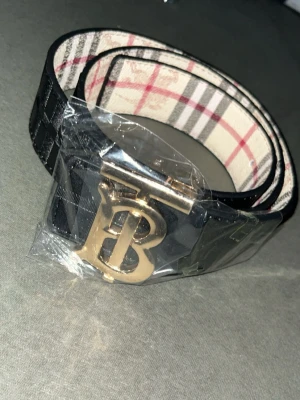 Burberry bälte med guldspänne - Snyggt Burberry bälte i svart skinn med klassiskt rutigt mönster på insidan och ett stort guldigt B-spänne framtill. Bältet har präglad logga på utsidan och ger en lyxig känsla till outfiten. Priset går diskutera 