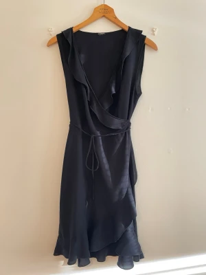 Svart omlottklänning med volang, xs, s - Snygg svart omlottklänning från Gina Tricot med volangdetaljer längs urringningen och nederkanten. Klänningen är ärmlös, har knyt i midjan och är tillverkad i ett lätt, mjukt material som ger ett fint fall.