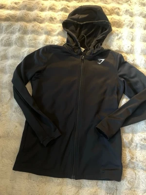 Svart hoodie från Gymshark med dragkedja - Svart hoodie från Gymshark med hel dragkedja och vit logga på bröstet. Tillverkad i ett mjukt och stretchigt material, perfekt för träning eller chill. Utrustad med huva och dragsko för extra komfort.