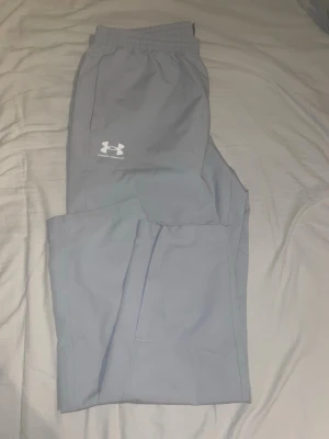 Ljusgrå träningsbyxor Under Armour - Snygga ljusgrå träningsbyxor från Under Armour med elastisk midja och logga på vänster lår. Perfekta för gymmet eller löprundan. Tillverkade i ett lätt och bekvämt material som andas.