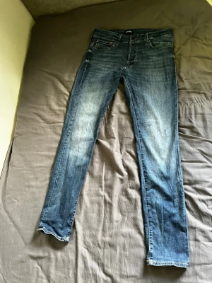 Blå raka jeans 32/34 - Säljer ett par klassiska blå jeans med raka ben och snygga slitningar. Jeansen har fem fickor, gylf med dragkedja och knapp samt kontrastsömmar. Perfekta för en avslappnad och stilren look.