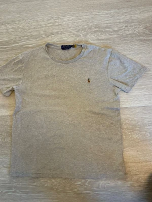 Beige t-shirt från Polo Ralph Lauren - Klassisk beige t-shirt från Polo Ralph Lauren med rund halsringning och korta ärmar. T-shirten har den ikoniska broderade polospelaren på bröstet och är tillverkad i mjuk bomull för en skön känsla. Perfekt basplagg med stilren design.