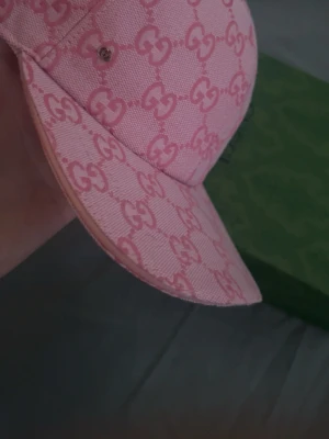Rosa gucci keps - Snygg rosa keps från Gucci med ikoniskt GG-monogram över hela kepsen. Kepsen har böjd skärm och är tillverkad i ett tygmaterial som känns både lyxigt och trendigt. Perfekt accessoar för att lyfta din outfit med en exklusiv touch. Boxen kommer inte med kepsen!