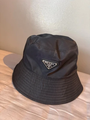 Svart bucket hat från Prada - Snygg svart bucket hat från Prada med klassisk trekantslogga framtill. Hatten är tillverkad i nylon och har en stilren, minimalistisk design som passar perfekt till streetwear. Märkeslapp och tvättråd finns på insidan.