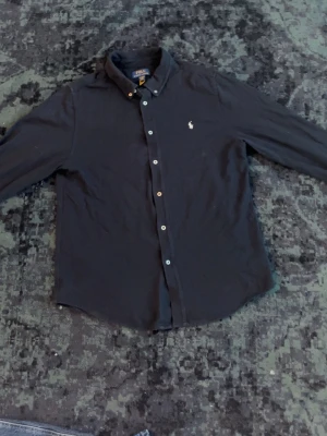 Blå Ralph lauren skjorta - Klassisk blå skjorta från Polo Ralph Lauren med lång ärm och button-down krage. Skjortan har vita knappar och den ikoniska broderade loggan på bröstet. Perfekt till jeans eller chinos för en stilren look.