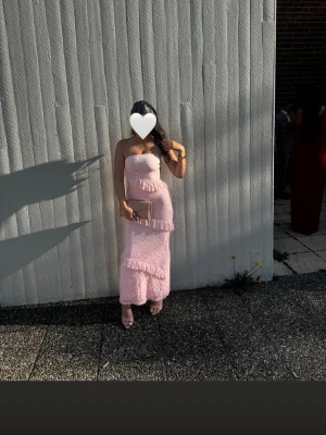 Ljusrosa axelbandslös långklänning - Säljer en ljusrosa axelbandslös långklänning med volangdetaljer och spetsmönster. Klänningen har en figurnära passform och är perfekt för sommarens speciella tillfällen. Klänningen är använd en kväll. Har en jätte litet hål i spetsen.(syns knappt)🩷