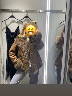 Brun pälsjacka mocka - Säljer en brun pälsjacka med stor, fluffig fuskpäls runt krage, ärmslut och längs öppningen. Jackan har en avslappnad passform och ger en cool vintagekänsla. 