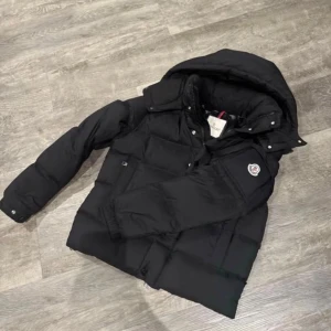 Svart Moncler pufferjacka med huva - Säljer en svart pufferjacka från Moncler med stor huva och klassisk logga på ärmen. Jackan har knappar och dragkedja framtill, samt två fickor på sidorna. Perfekt för kalla dagar och har en riktigt schysst, boxig passform.