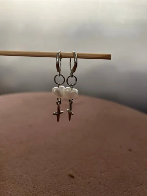 Hemma gjorda Silverörhängen med hjärtpärlor och stjärnor - Säljer ett par hemma gjorda snygga örhängen i silverfärg med små vita hjärtformade pärlor och hängande stjärnberlocker. Perfekta för dig som gillar unika detaljer och vill sticka ut lite extra. Passar till alla tillfällen och ger en söt touch till din stil.