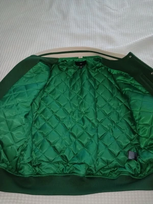 Grön quiltad bomberjacka H&M - Snygg grön bomberjacka från H&M med quiltat foder och ribbade muddar. Jackan har en klassisk bombermodell och är tillverkad i polyester med lite elastan för extra komfort. Perfekt för dig som vill sticka ut med en färgstark look.