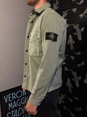 Grön overshirt från Stone Island - Säljer en snygg ljusgrön overshirt från Stone Island med klassisk patch på vänster ärm. Skjortan har bröstficka med knapp, svarta knappar framtill och är tillverkad i bomull. Perfekt för lager på lager och en clean streetwear-look.