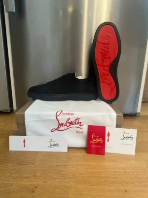 Svarta sneakers från Christian Louboutin - Stilrena svarta sneakers från Christian Louboutin med ikonisk röd sula och diskret logga på sidan. Skorna har en låg profil och är tillverkade i mocka med klassiska svarta skosnören. Perfekta för dig som vill ha en lyxig och tidlös sneaker med statement-detaljer.