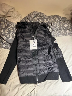 Svart pufferjacka från Moncler - Stilren svart pufferjacka från Moncler med quiltad front, stickade ärmar och huva. Jackan har dragkedja, sidofickor med knappar och Moncler-logga på ärmen. Perfekt för kalla dagar och ger en snygg, sportig look.
