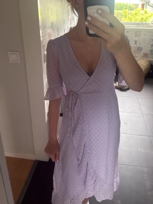 Ljuslila omlottklänning med prickar - Söt ljuslila omlottklänning med volangdetaljer. Klänningen har v-ringning, knyt i midjan och trekvartsärm med volang vid ärmslut. Perfekt till sommaren💕