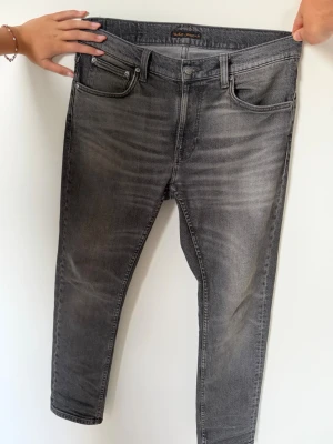 Grå slim fit jeans från Nudie Jeans - Snygga grå slim fit jeans från Nudie Jeans med klassisk femficksmodell och diskret tvättad look. Jeansen har normal midja, dragkedjegylf och är tillverkade i mjukt denimtyg. Perfekta för en avslappnad och trendig stil.