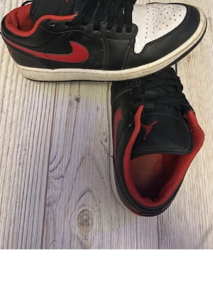 Svarta och röda Air Jordan 1 Low - Säljer ett par Air Jordan 1 Low sneakers i svart och rött med vit tåbox. Skorna har klassisk röd swoosh, röd insida och ikoniska Air Jordan-loggor på hälen. Tillverkade i läder med snörning framtill. Perfekta för dig som gillar streetwear och snygg design. 