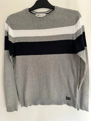 Grå stickad tröja med ränder H&M - Snygg grå stickad tröja från H&M med breda vita och mörkblå ränder över bröstet. Tröjan har rund halsringning, ribbade muddar och en liten NYCME-detalj nertill. Perfekt för dig som gillar enkel och stilren design.