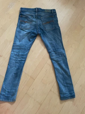 Nudie jeans tilted tor  - Snygga blå  nudie jeans med klassisk femficksdesign och slitningar på benen. Modellen är tilted Tor. Jeansen har raka ben och detaljerade sömmar på bakfickorna. Tillverkade i denim, perfekta för en avslappnad stil.