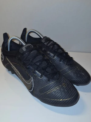 Nike svarta fotbollsskor med guld - Snygga svarta fotbollsskor från Nike med gulddetaljer och vita loggor på sidan. Skorna har snörning och en låg profil för bra bollkontroll. Ovandelen är i slitstarkt syntetmaterial och sulan har dobbar för bra grepp på planen.
