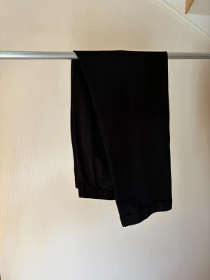 Svarta kostymbyxor med pressveck - Snygga svarta kostymbyxor med klassiskt pressveck och uppvikta benslut. Byxorna har hög midja, sidofickor och stängs med knapp och dragkedja. Perfekta för en stilren och clean look. Materialet ser ut att vara en mjuk och lätt vävd kvalitet.