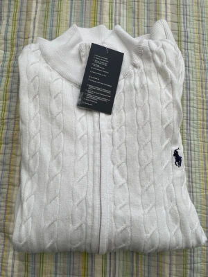 Vit kabelstickad tröja från Polo Ralph Lauren - Kabelstickad vit tröja med dragkedja från Polo Ralph Lauren. Tröjan har ribbade muddar och den klassiska broderade loggan i marinblått på bröstet. Perfekt för dig som gillar en clean och tidlös look.