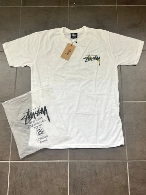 Vit Stussy t-shirt med camologga - Säljer en vit t-shirt från Stussy med klassisk logga i camomönster på bröstet. T-shirten har rund hals och korta ärmar. Perfekt för dig som gillar streetwear och vill ha något stilrent men ändå med en twist. 3 pyttesmå ljusröda fläckar i mitten som går bort med tvätt