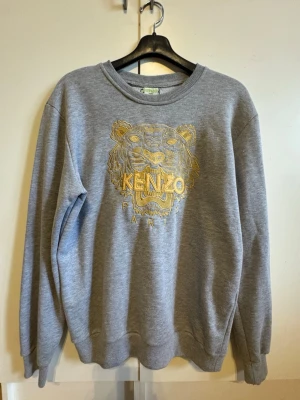 Grå Kenzo sweatshirt med broderat tigertryck i guld - Snygg grå sweatshirt från Kenzo med ikoniskt broderat tigerhuvud och Kenzo Paris-logga i guld på bröstet. Tröjan har rund halsringning, ribbade muddar och är tillverkad i mjukt material. Perfekt för en trendig och avslappnad stil.