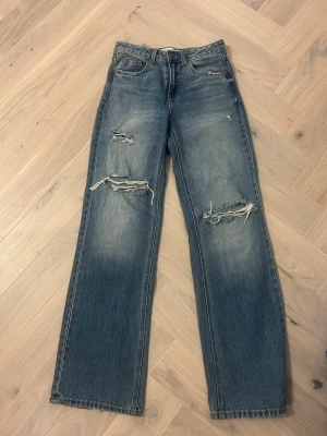 Blå slitna raka jeans Stradivarius - Ett par blå raka jeans från Stradivarius med slitningar och hål på båda benen. Jeansen har klassisk femficksmodell, hög midja och är gjorda i denim. Perfekta för en avslappnad och trendig look.