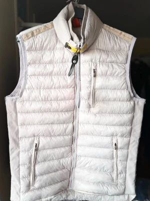 Vit dunväst med hög krage - Snygg vit dunväst med quiltad design och hög krage. Västen har dragkedja framtill, två sidofickor med dragkedja och en bröstficka. Detaljer i gult och svart vid halsen ger en cool touch. Perfekt för lager-på-lager och streetwear.