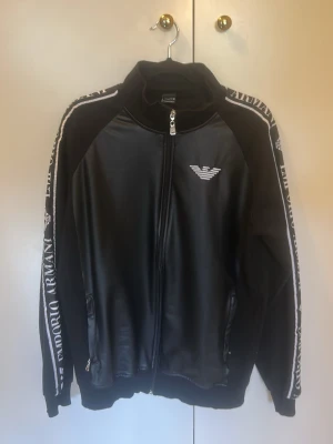 Svart skinjacka från Armani - Snygg svart skinjacka från Emporio Armani med dragkedja framtill och vita logotyper på bröstet och längs ärmarna. Jackan har hög krage, två fickor med dragkedja och vita detaljer längs sidorna. Perfekt för sport eller streetwear.